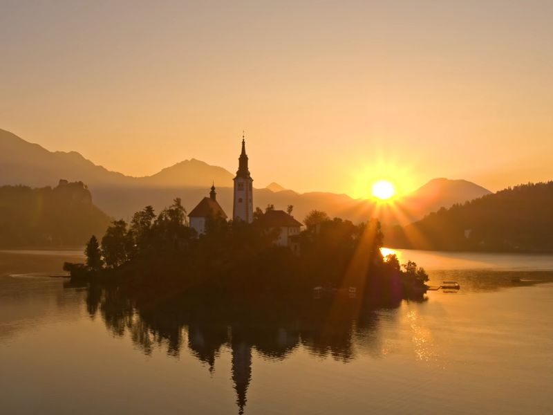 Zalazak sunca na Jezeru Bled u Sloveniji, sa ostrvom u sredini na kojem se nalazi crkva, okruženo mirnom vodom i planinskim pejzažom u pozadini.
