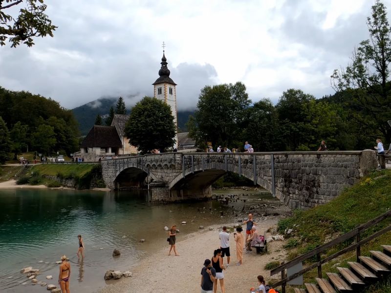 Ljudi uživaju pored jezera Bohinj, sa kamenitim mostom i crkvom u pozadini, okruženi zelenilom i planinama pod oblačnim nebom.